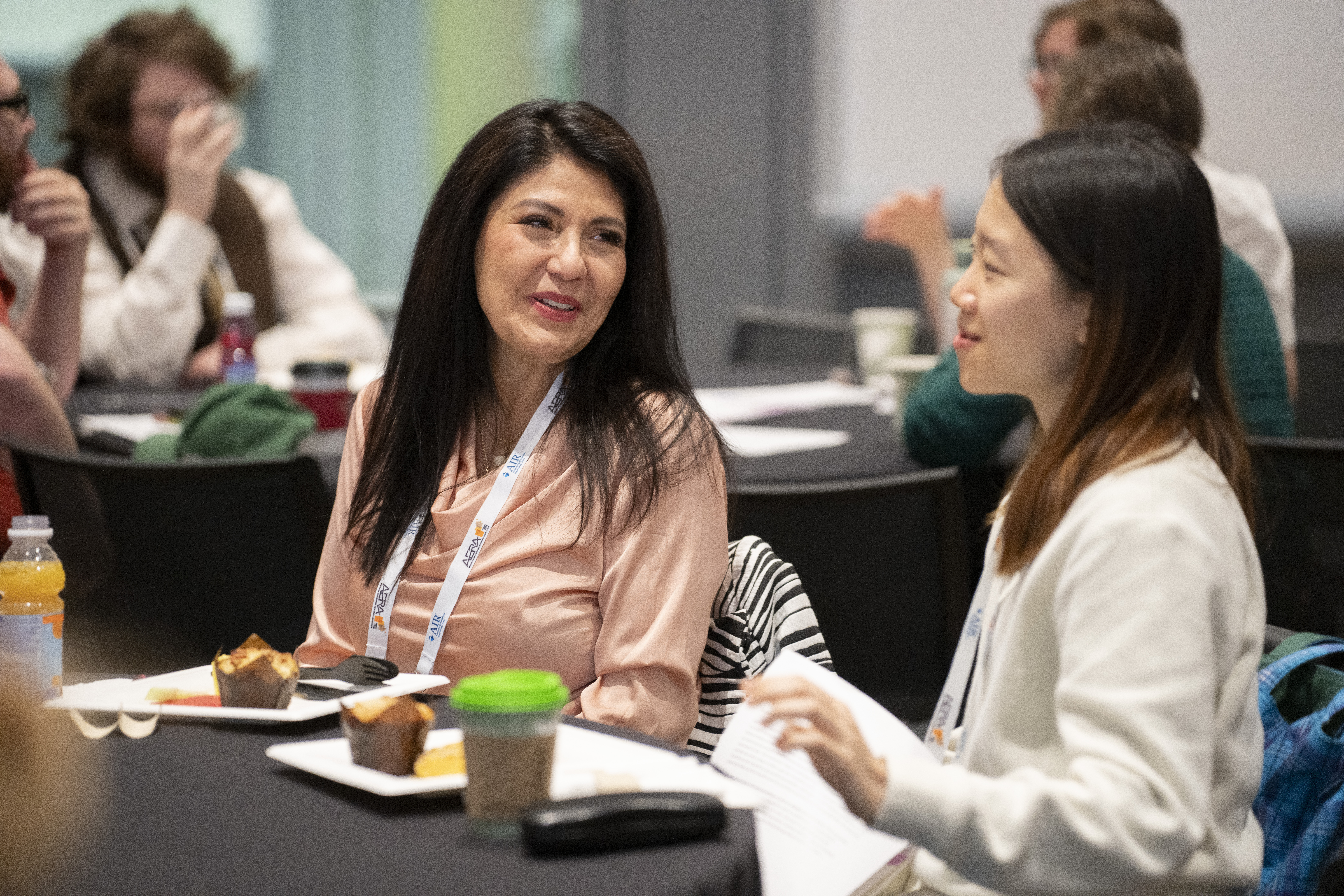 Two women at a table during a conference. They have coffee in front of them on the table.
