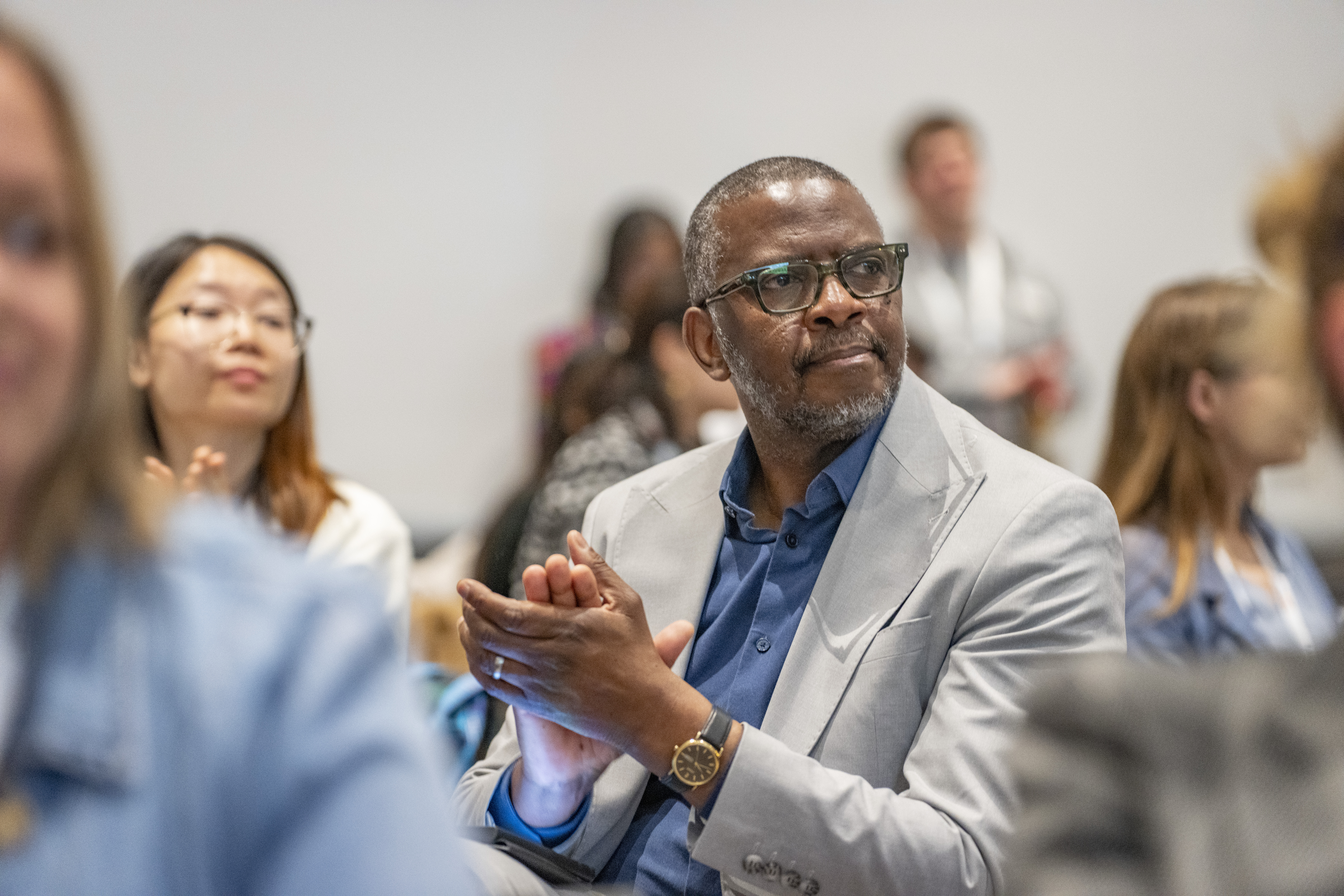 A man sits at a table surrounded by conference attendees. He is looking to the side and applauding the speaker.
