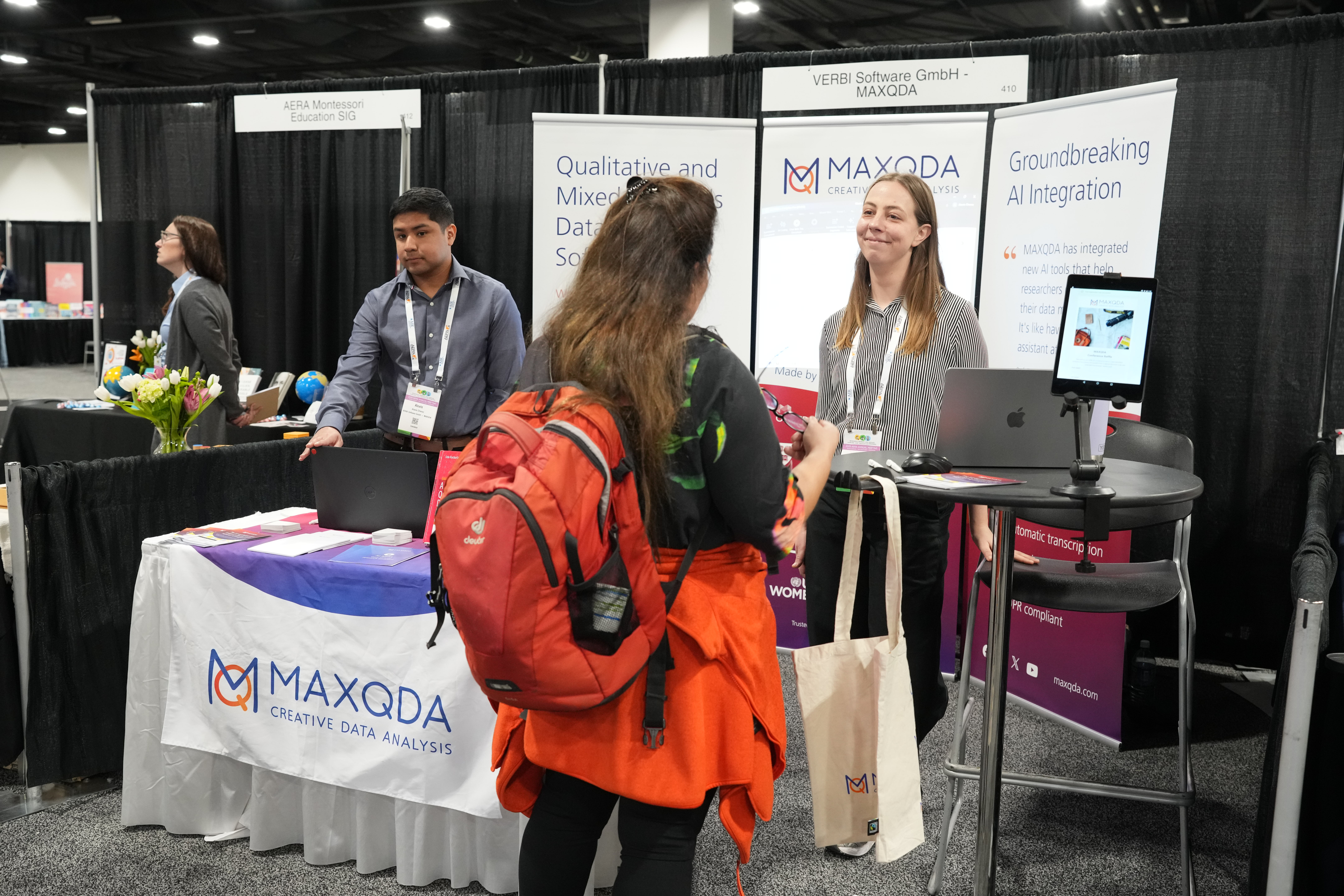 A few people stand behind a booth at a conference advertising their products. One of the people is speaking with an attendee who is not facing the camera.