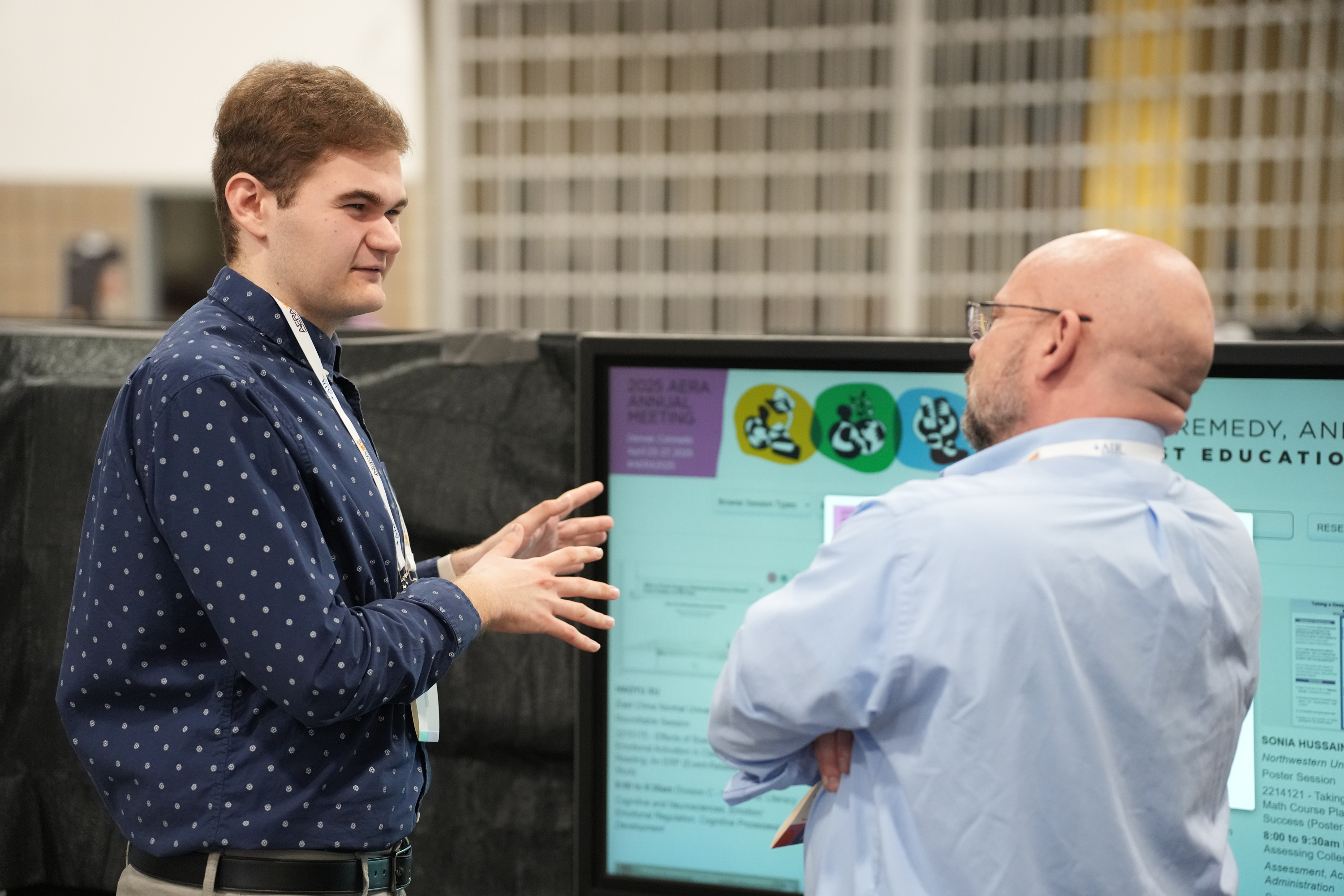 Two men are speaking in front of a poster. One man is facing away from the camera while the other man is facing toward the poster and gesturing at it.