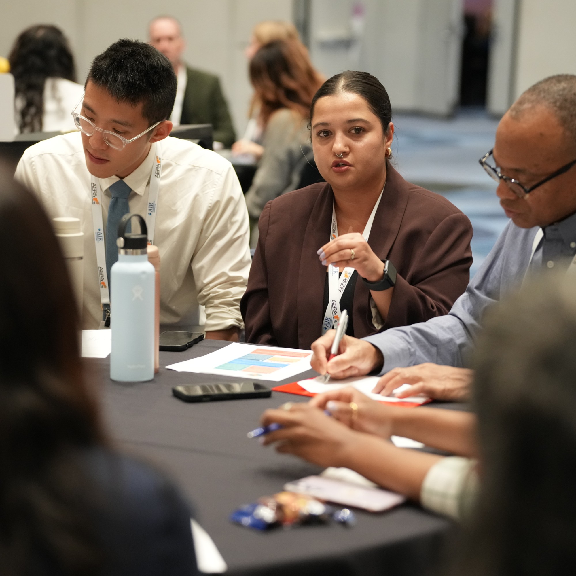 A group of attendees at a roundtable session. One woman is talking with her hands and the others are looking down to their notebooks and writing
