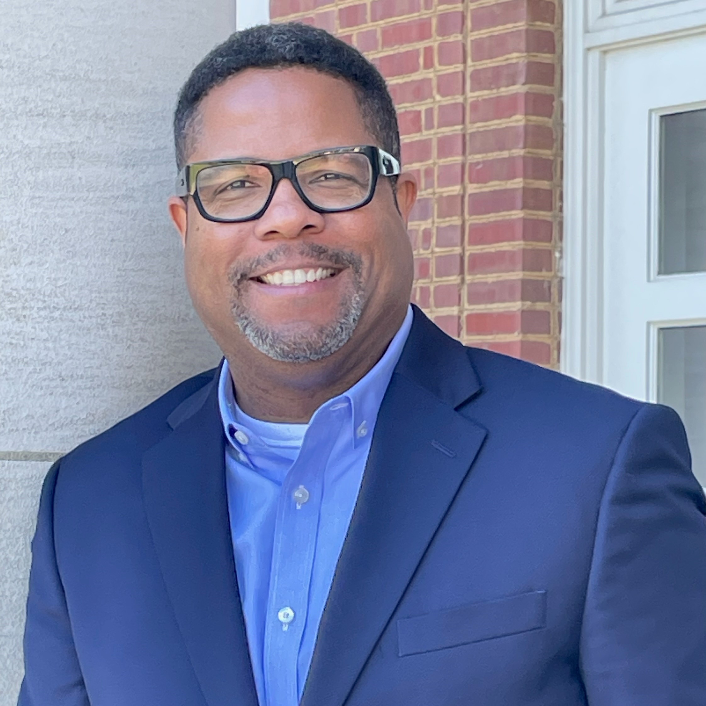 zoomed in color image of Rich Milner. He is wearing a suit and button up shirt with glasses, standing against a grey and brick background