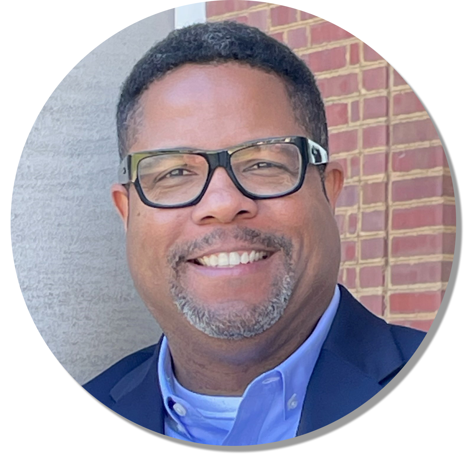 zoomed in color image of Rich Milner. He is wearing a suit and button up shirt with glasses, standing against a grey and brick background